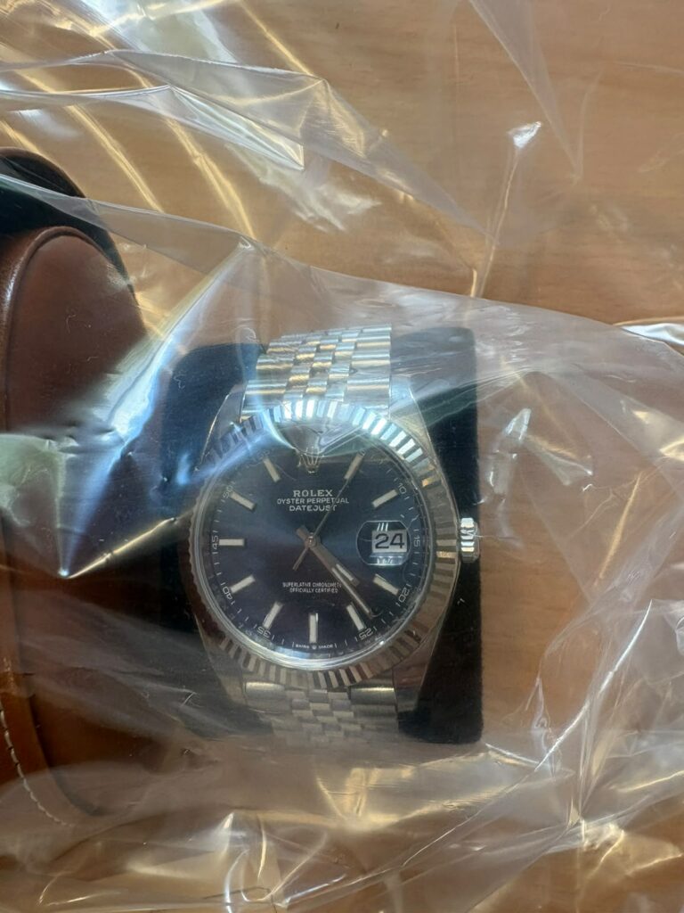 Montre à cadran sombre et bracelet métallique sous un sachet plastique transparent.