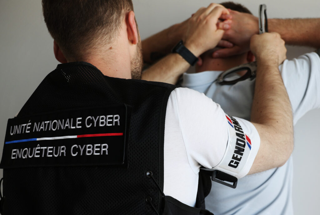 Enquêteur portant un gilet Unité nationale cyber et un brassard Gendarmerie, tenant des menottes derrière une personne.